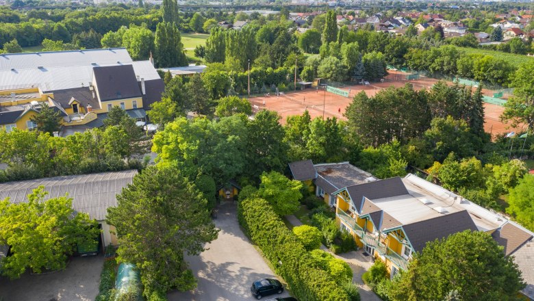 Luftaufnahme eines Tennisplatzes mit umliegenden Geb&auml;uden und B&auml;umen.