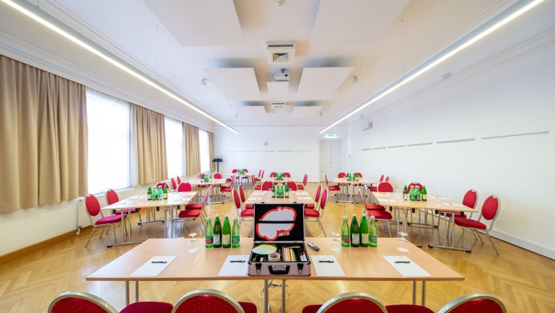 Ein heller Seminarraum mit roten Stühlen und Tischen, auf denen Wasserflaschen und Schreibmaterialien stehen.