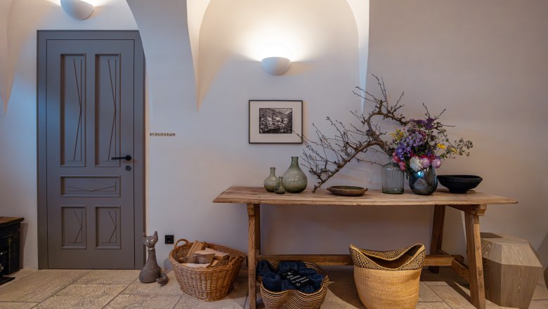 Innenraum mit Holztisch, Vasen, Blumen und K&ouml;rben. T&uuml;r mit Aufschrift 'Schuhraum'.