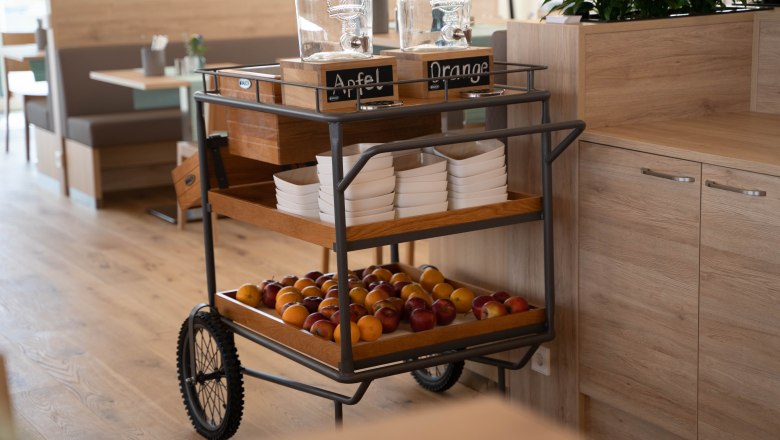 Frühstücksbereich mit einem Servierwagen voller Obst und Getränkespendern.