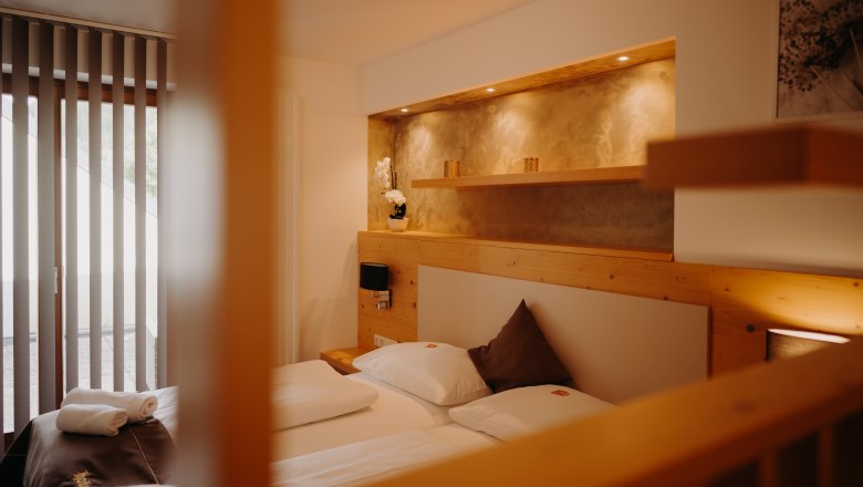 Gem&uuml;tliches Appartement mit Doppelbett und Balkont&uuml;r. Die Wand ist zum Teil mit hellem Holz verkleidet, hinter dem Bett ist eine in die Wand eingelassene Nische mit Ablagefl&auml;che und Spots, in einem erdigen Gold gestrichen.