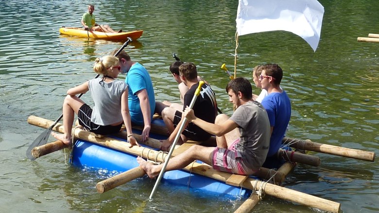 Gruppe von Menschen auf einem selbstgebauten Floß auf einem See, mit einer weißen Flagge und einem Kajakfahrer im Hintergrund.
