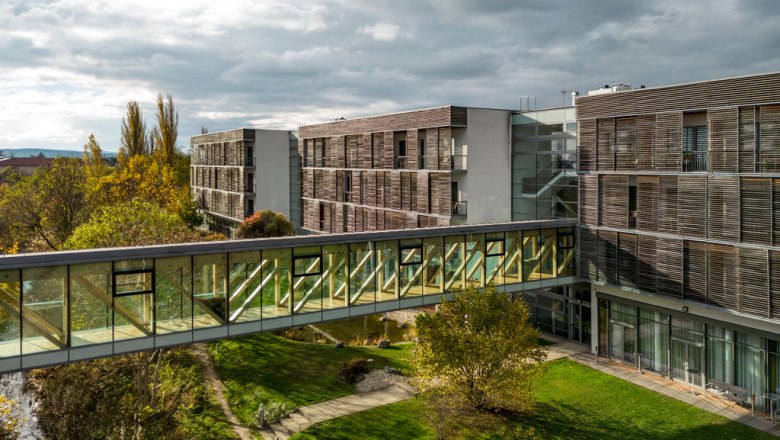 Glas&uuml;berdachter &Uuml;bergang zwischen Therme Laa und Hotelgeb&auml;ude, umgeben von gr&uuml;ner Landschaft.