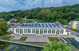 Luftaufnahme einer alten Fabrik mit Solarpanelen auf dem Dach, umgeben von B&auml;umen und einem Teich.