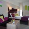 Modern eingerichtetes Hotelzimmer mit Doppelbett, Sofa und Dekoration in Lila- und Gr&uuml;nt&ouml;nen.