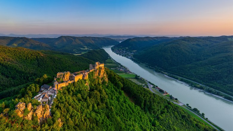 Luftaufnahme der Burgruine Aggstein an der Donau, umgeben von gr&uuml;nen H&uuml;geln und W&auml;ldern bei Sonnenuntergang.