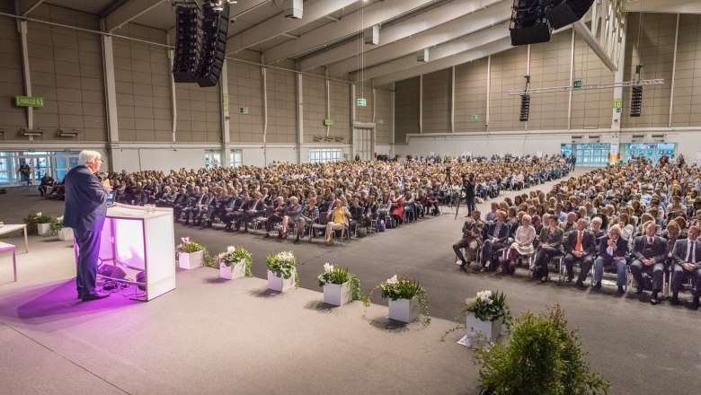 Ein Redner spricht vor einem gro&szlig;en Publikum in einer Messehalle.
