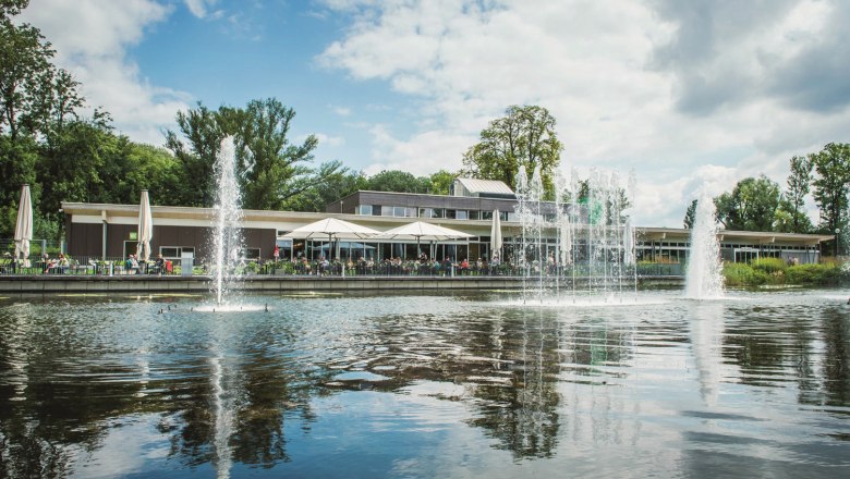 Ein modernes Gebäude mit Terrasse am Wasser, umgeben von Bäumen und Springbrunnen.