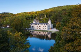 Schloss Hernstein mit Spiegelung im Wasser, umgeben von Bäumen und Hügeln.