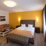 Hotelzimmer mit Doppelbett, gelber Wand, Sofa, Tisch und Vorh&auml;ngen.