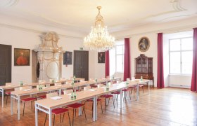 Eleganter Seminarraum mit Kronleuchter, Holzboden und Porträts an den Wänden.