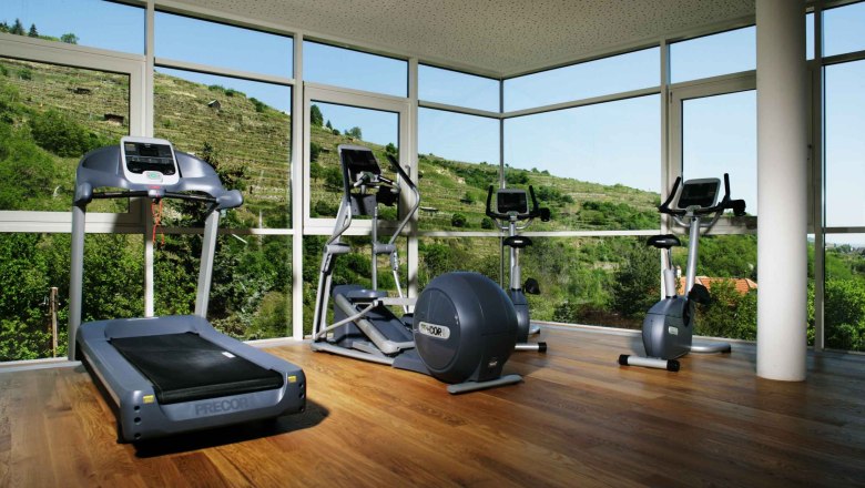 Fitnessraum mit Laufband, Crosstrainer und Heimtrainer vor gro&szlig;en Fenstern mit Blick auf gr&uuml;ne H&uuml;gel.