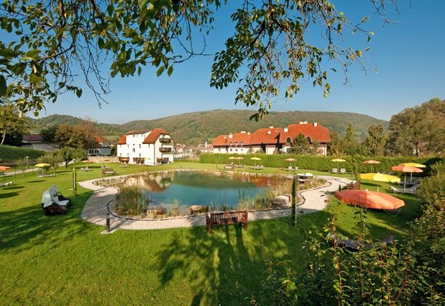 Gartenanlage mit Teich, Liegest&uuml;hlen und Sonnenschirmen, umgeben von Geb&auml;uden und H&uuml;geln.
