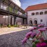 Ein Innenhof mit Kopfsteinpflaster, Tischen und St&uuml;hlen, umgeben von Geb&auml;uden mit Arkaden und einem modernen Balkon.
