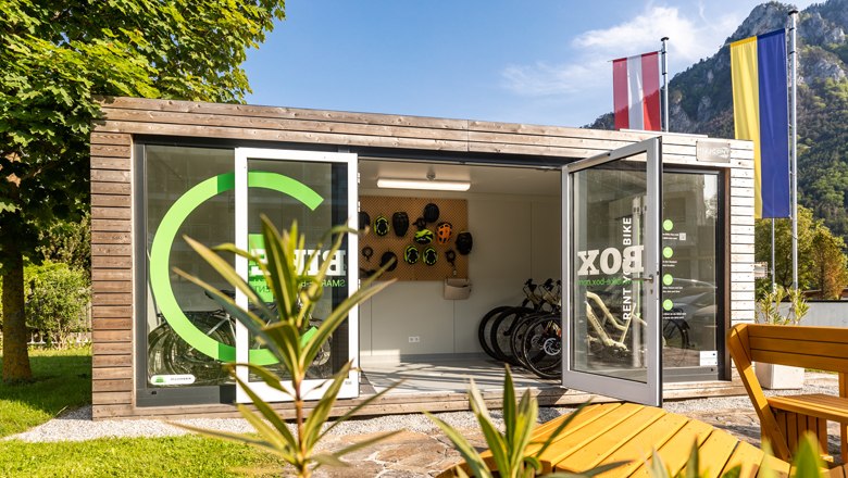 Eine moderne Fahrradbox mit ge&ouml;ffneten T&uuml;ren, Fahrr&auml;dern und Helmen, umgeben von Natur und Flaggen.