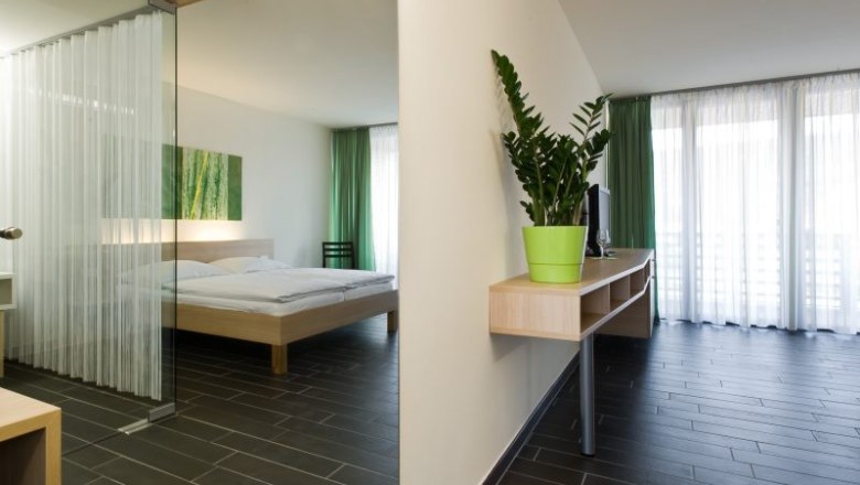 Modernes Hotelzimmer mit Doppelbett, gr&uuml;nem Vorhang und Zimmerpflanze.
