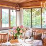 Elegantes Restaurant mit gedeckten Tischen, Blumen und Blick auf die Natur durch gro&szlig;e Fenster.