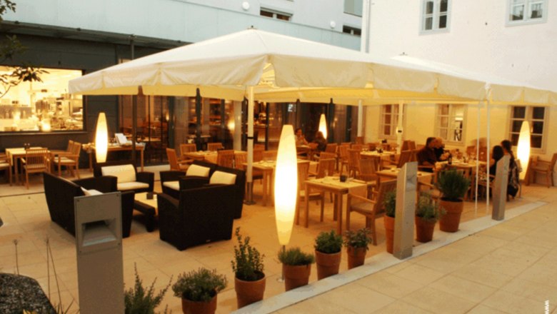 Ein beleuchteter Gastgarten mit Tischen, St&uuml;hlen und Pflanzen in T&ouml;pfen, umgeben von Geb&auml;uden.