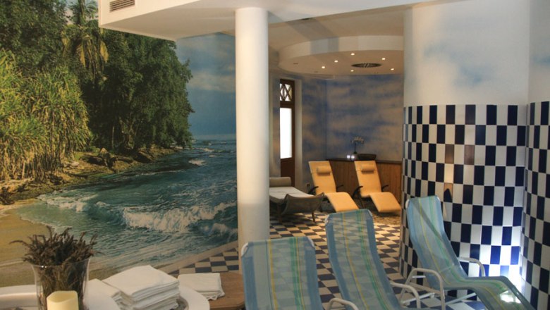 Ein Saunabereich mit Liegest&uuml;hlen, einem Wandbild von einem Strand und blau-wei&szlig; karierten Fliesen.