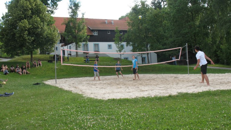 Menschen spielen Volleyball auf einem Sandplatz im Freien, umgeben von B&auml;umen und einem Geb&auml;ude im Hintergrund.