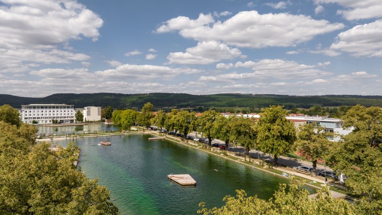 Luftaufnahme eines Sees mit umliegenden Geb&auml;uden und B&auml;umen unter blauem Himmel.