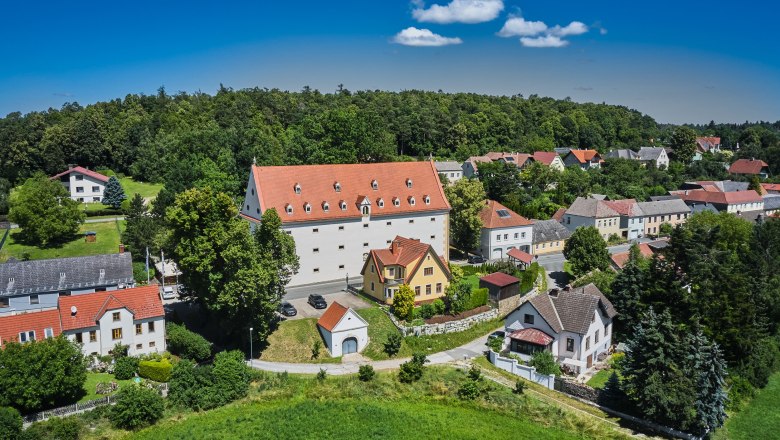 Luftaufnahme eines Dorfes mit einem großen weißen Gebäude mit rotem Dach, umgeben von kleineren Häusern und viel Grün.