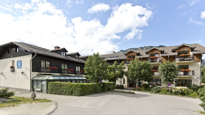 Ein traditionelles Gasthaus mit Holzbalkonen und Blumenkästen, umgeben von Bäumen und Bergen im Hintergrund.