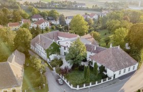 Luftaufnahme eines historischen Gebäudekomplexes mit Innenhof, umgeben von Bäumen und weiteren Häusern in einer ländlichen Gegend.