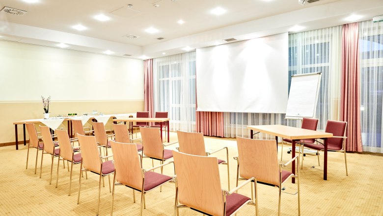 Ein heller Seminarraum mit Stühlen, Tischen, einem Flipchart und einem Projektionsschirm.