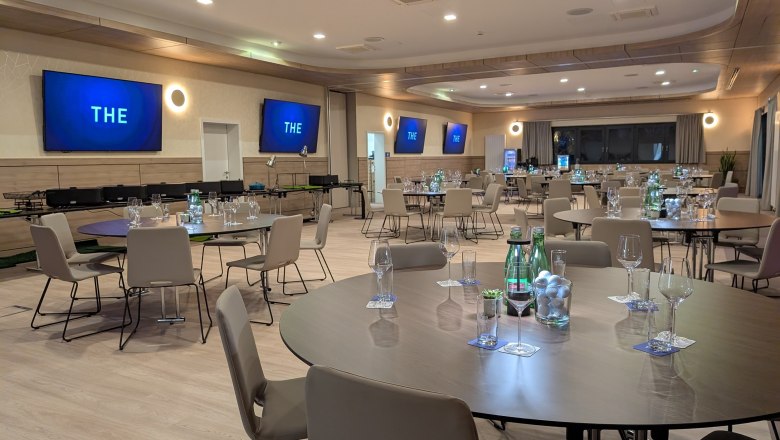 Moderner Seminarraum mit runden Tischen, Stühlen und Bildschirmen an den Wänden.