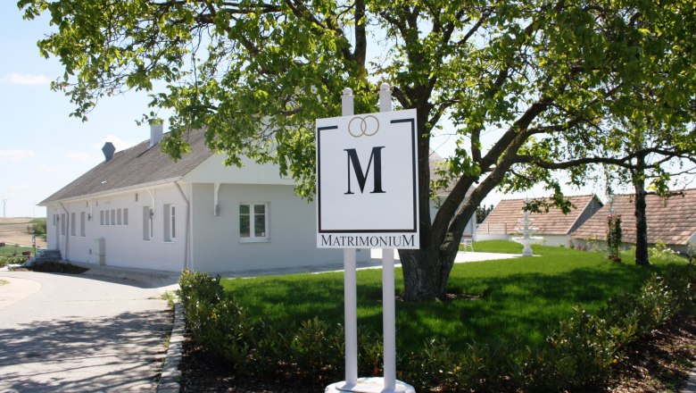 Ein weißes Gebäude mit einem Schild "Matrimonium" im Vordergrund, umgeben von grüner Landschaft.