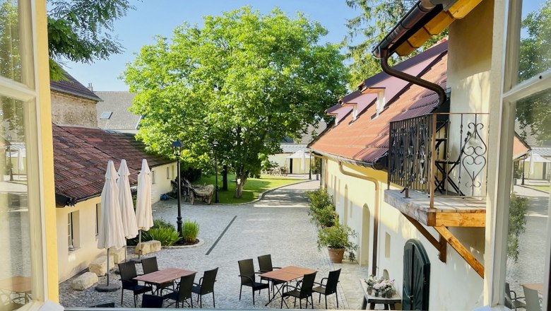 Blick aus einem Fenster auf einen Innenhof mit Tischen, Stühlen und Sonnenschirmen, umgeben von Gebäuden und Bäumen.