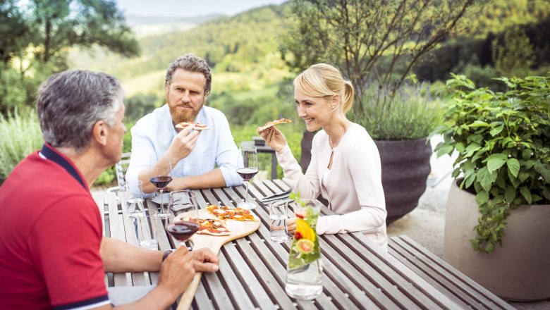 Drei Personen essen Pizza und trinken Wein an einem Holztisch im Freien mit Blick auf eine grüne Landschaft.