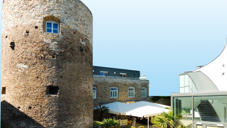 Historischer Turm neben modernem Geb&auml;ude mit Terrasse und Sonnenschirmen.