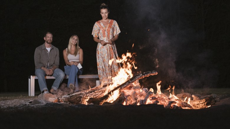 Drei Personen sitzen und stehen um ein Lagerfeuer in der Nacht.