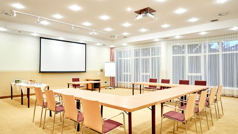 Ein heller Seminarraum mit U-förmiger Tischanordnung, Leinwand, Flipchart und Getränketisch.