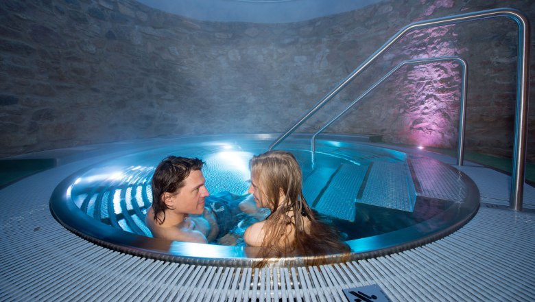 Ein Paar entspannt in einem runden Whirlpool mit blauer Beleuchtung und Steinw&auml;nden.