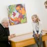 Zwei Pater sprechen mit zwei Kindern in einer Galerie vor einem bunten Gem&auml;lde.