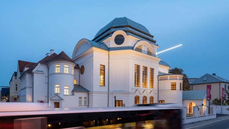 Ehemalige Synagoge in St. P&ouml;lten bei D&auml;mmerung mit beleuchteten Fenstern und vorbeifahrendem Bus.