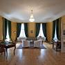 Elegantes Zimmer mit Holzboden, gr&uuml;nen Vorh&auml;ngen, zwei Sesseln, einem Sofa und einem Schreibtisch. Ein Kronleuchter h&auml;ngt von der Decke.