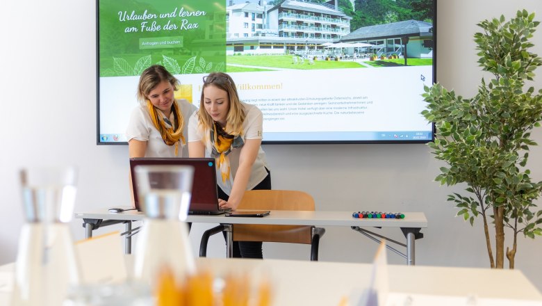 Zwei Frauen arbeiten an einem Laptop vor einem großen Bildschirm mit einer Hotel-Website. Im Vordergrund sind Gläser und Stifte zu sehen.