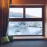 Blick aus einem Holzfenster auf eine verschneite Landschaft mit Bergen im Hintergrund.