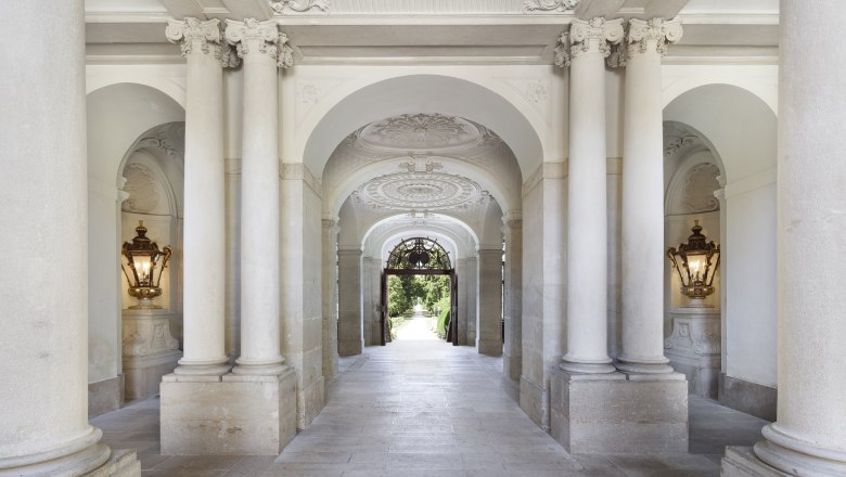 Eleganter, mit Säulen gesäumter Durchgang im Schloss Eckartsau mit verzierten Decken und Wandleuchten.