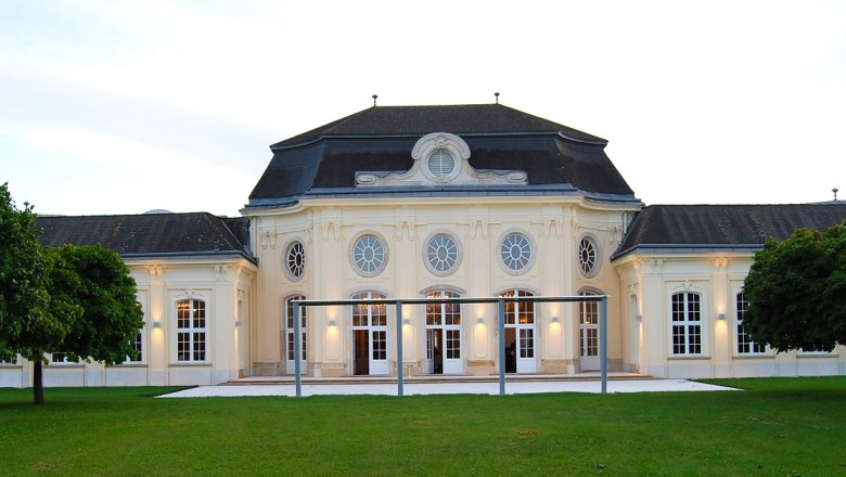 Historisches Gebäude mit symmetrischer Fassade und großen Fenstern, umgeben von Grünflächen.
