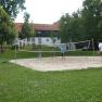 Menschen spielen Volleyball auf einem Sandplatz im Freien, umgeben von B&auml;umen und einem Geb&auml;ude im Hintergrund.
