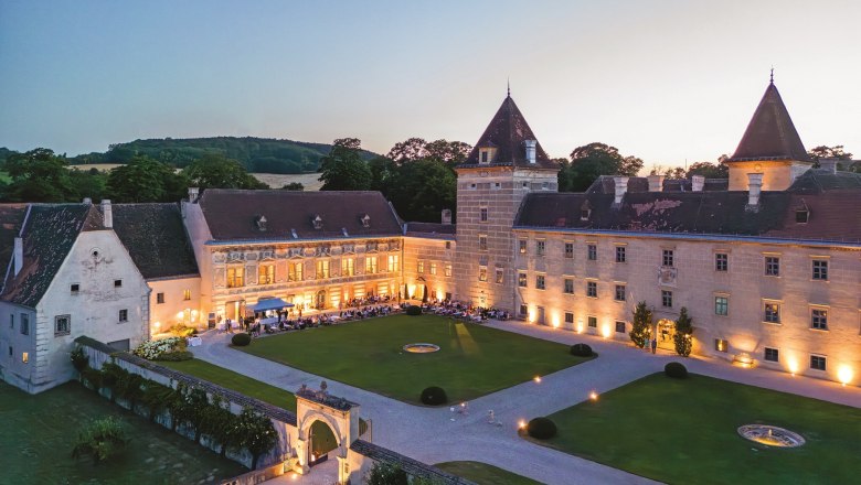 Luftaufnahme von Schloss Walpersdorf bei Dämmerung, beleuchtet mit Gästen bei einem Gartenfest im Innenhof.