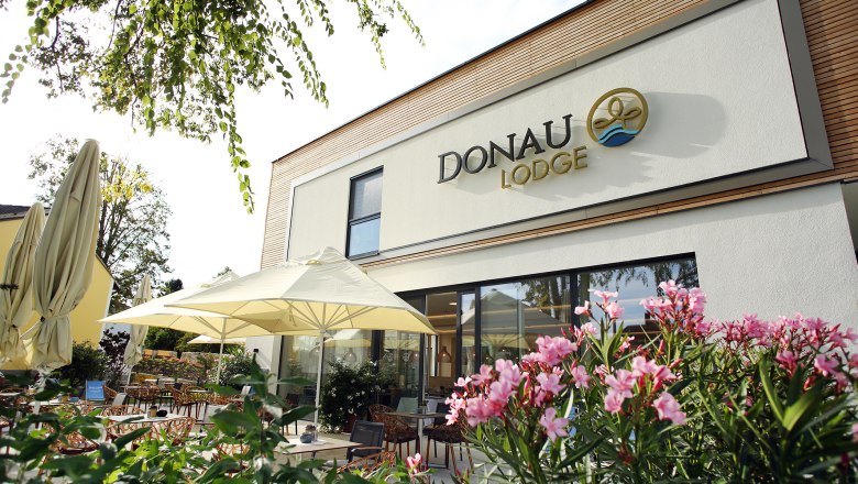 Au&szlig;enansicht der Donaulodge mit Terrasse, Sonnenschirmen und bl&uuml;henden Pflanzen.
