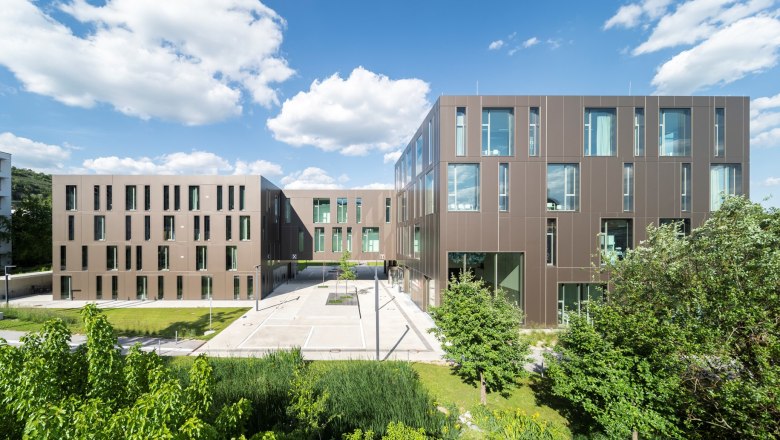 Moderne Universitätsgebäude mit brauner Fassade und großen Fenstern, umgeben von grüner Vegetation unter blauem Himmel.