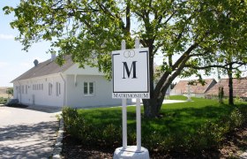 Ein weißes Gebäude mit einem Schild "Matrimonium" im Vordergrund, umgeben von grüner Landschaft.
