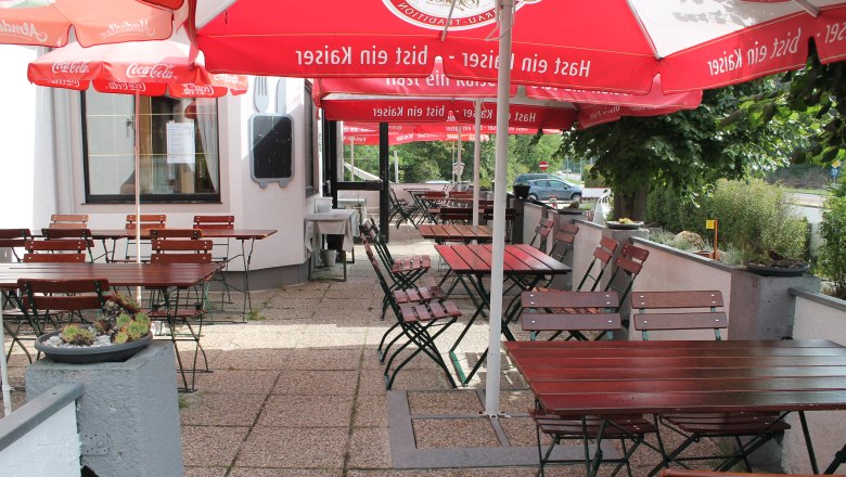 Ein leerer Gastgarten mit Holztischen und -st&uuml;hlen unter roten Sonnenschirmen.